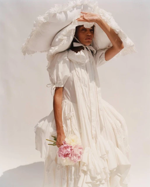 Person in ruffled white dress and oversized hat shields their eyes while holding flowers against a plain white background.