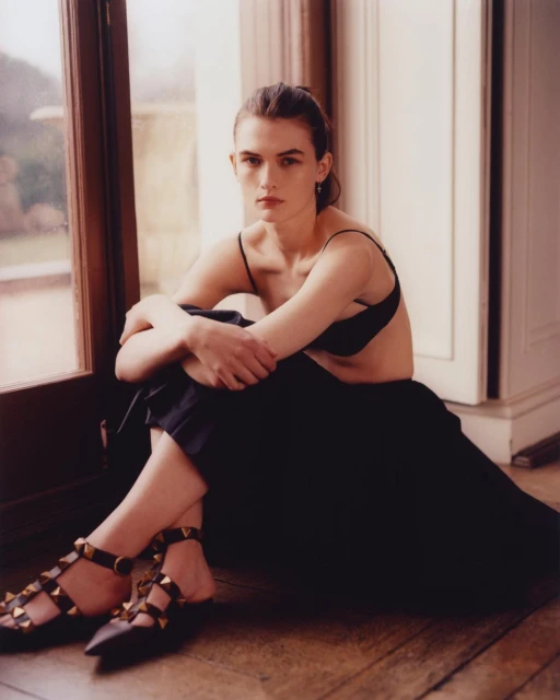 Portrait of a brunette woman with dark skirt and studded shoes sitting by a window in a sparse room with a glimpse of a garden.
