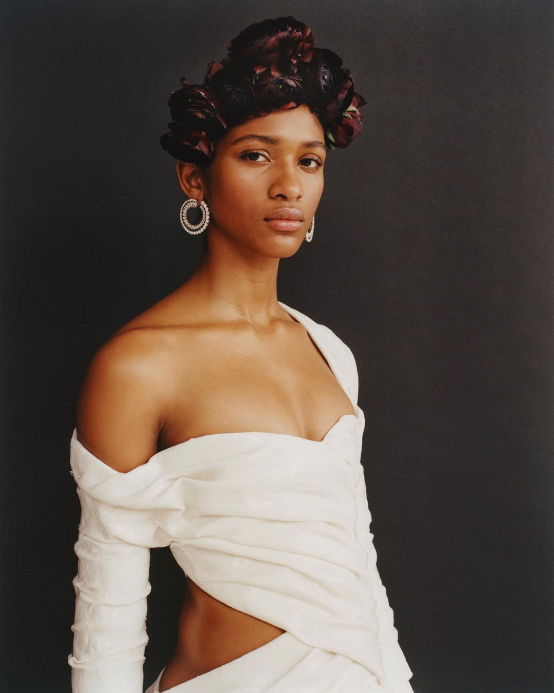 A portrait shot of a woman with dark hair, wearing a light-colored dress and statement earrings, against a dark grey backdrop in a studio.