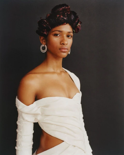 A portrait shot of a woman with dark hair, wearing a light-colored dress and statement earrings, against a dark grey backdrop in a studio.