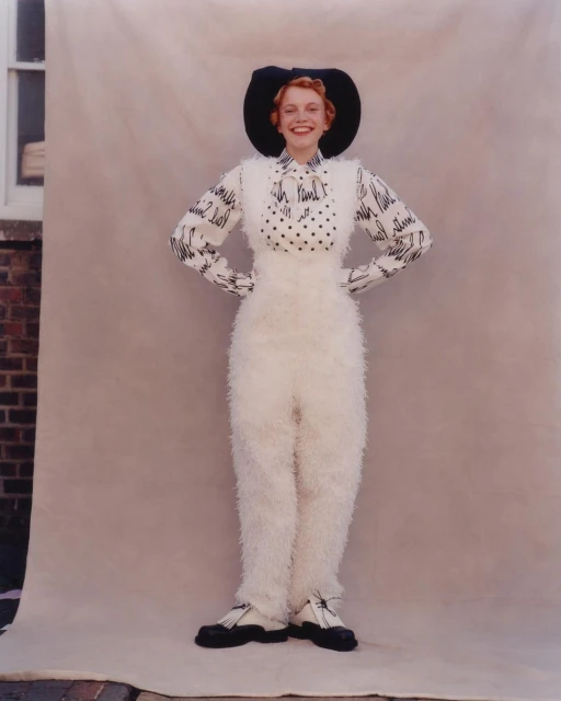 Full-length portrait of a smiling woman in a patterned shirt, fuzzy white overalls, hat, and black shoes, standing in front of a backdrop.