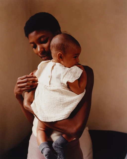 African American mother affectionately embraces her baby indoors. The baby wears a simple white dress and gray socks in soft, natural light.