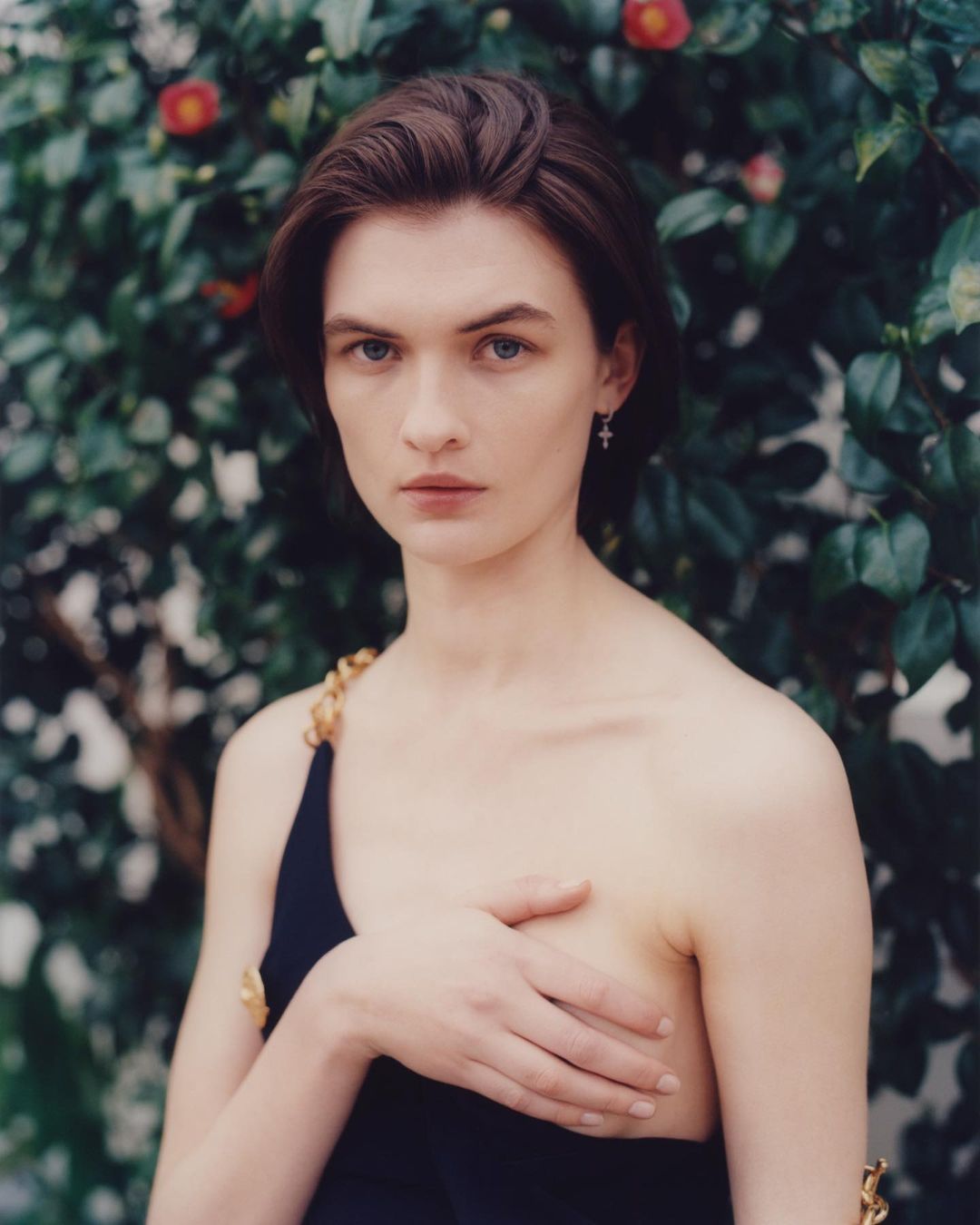 Elegant woman in a dark sleeveless dress poses in front of green foliage with red flowers. She is wearing earrings and has short brown hair.