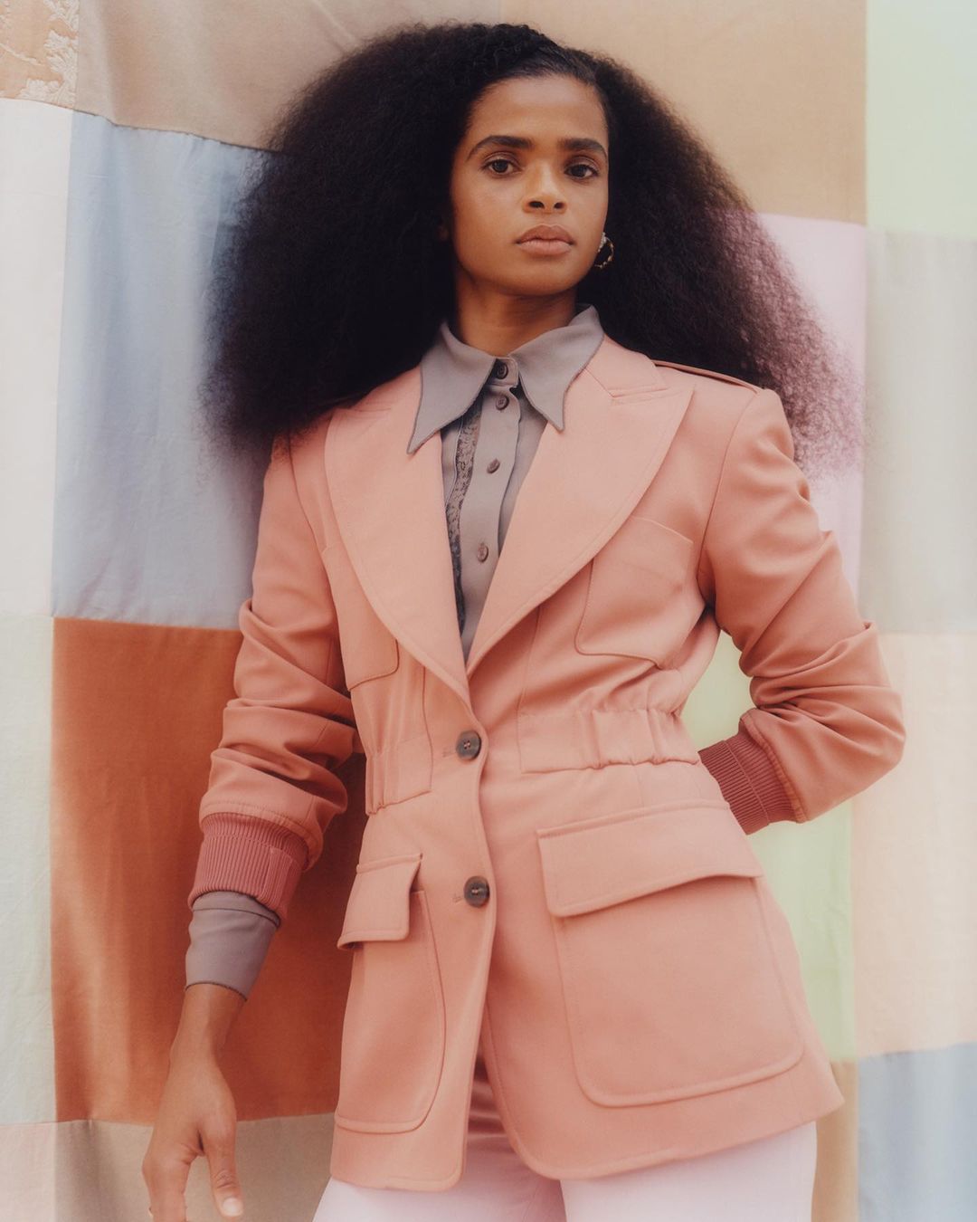 Portrait of a woman with voluminous dark hair wearing a salmon-colored blazer with patch pockets against a pastel color-blocked backdrop.