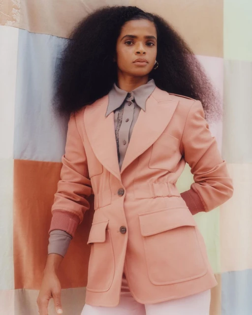 Portrait of a woman with voluminous dark hair wearing a salmon-colored blazer with patch pockets against a pastel color-blocked backdrop.