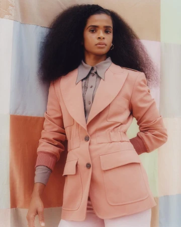 Portrait of a woman with voluminous dark hair wearing a salmon-colored blazer with patch pockets against a pastel color-blocked backdrop.