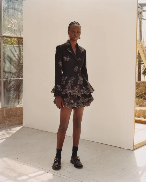 Full-length portrait of a Black female fashion model posing in a black blazer dress with floral embroidery in a light-filled indoor setting.