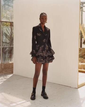 Full-length portrait of a Black female fashion model posing in a black blazer dress with floral embroidery in a light-filled indoor setting.