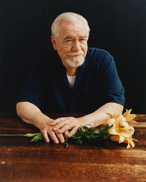 Portrait of actor Brian Cox with a slight smile, seated at a wooden table with a yellow lily, set against a dark backdrop.