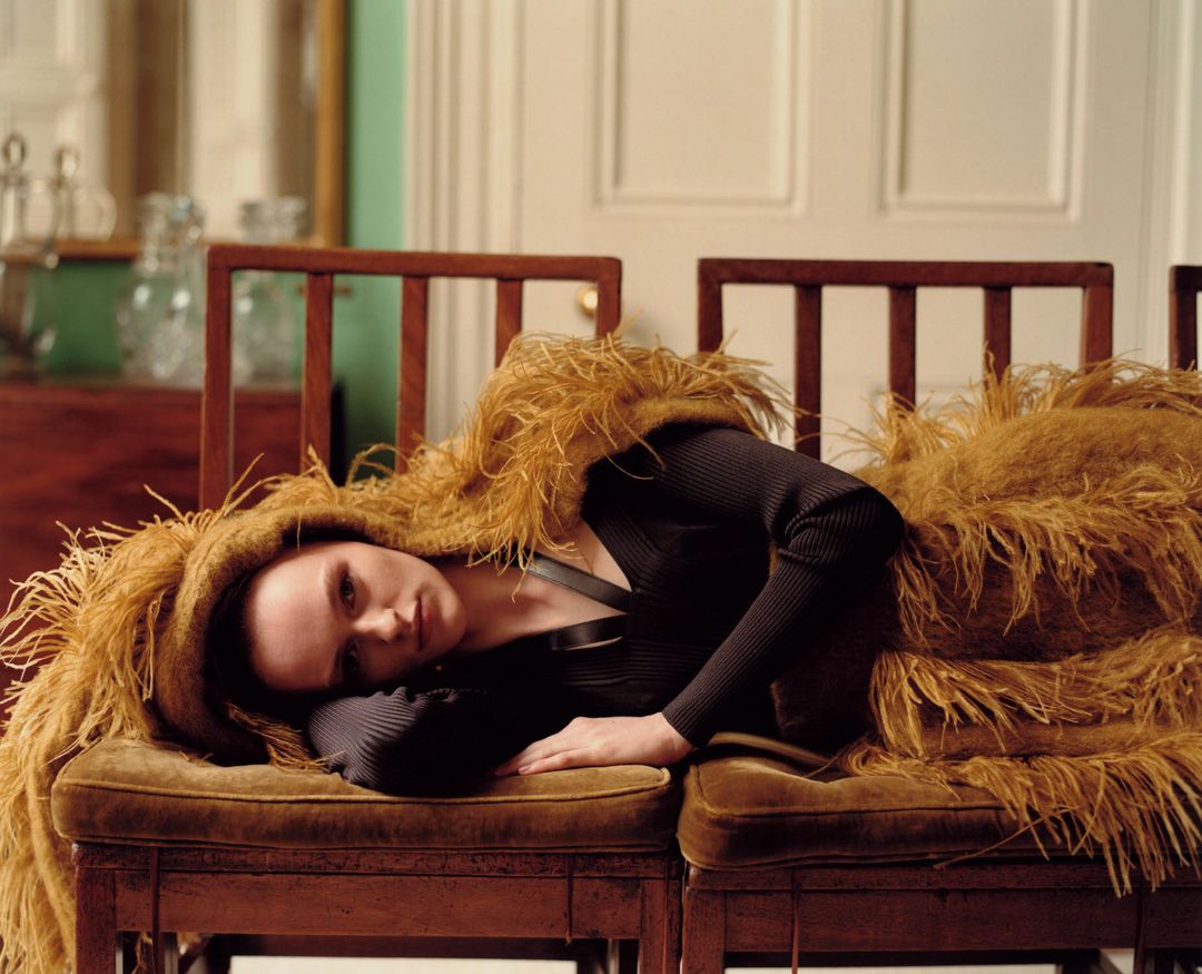 Brunette woman in black top and orange feathered wrap reclines on brown cushioned chair indoors, illuminated by natural light.