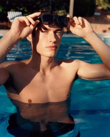 Shirtless man adjusting black sunglasses while standing waist-deep in a bright blue outdoor swimming pool