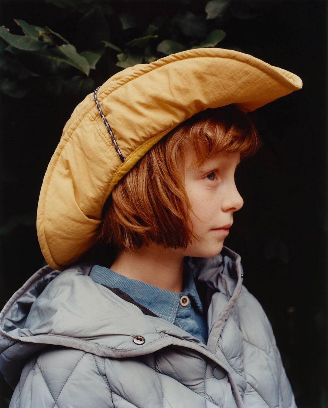 Portrait of a child with red hair wearing a yellow hat and grey puffer jacket, looking left against a dark green leafy background.