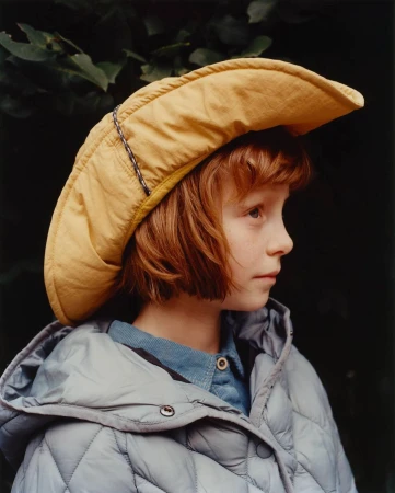 Portrait of a child with red hair wearing a yellow hat and grey puffer jacket, looking left against a dark green leafy background.