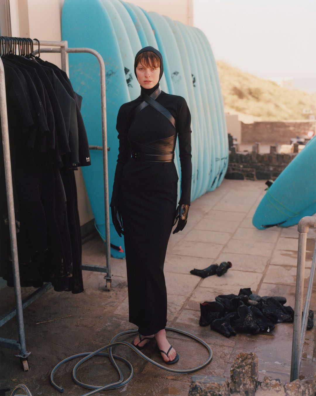 A striking woman in a black dress and head covering stands on a tiled surface, surfboards behind her, wetsuits hanging on a rack.