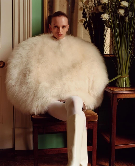 A woman in a white, feathery coat sits on a wooden stool in a studio with a cream-colored door and a bouquet of white flowers.