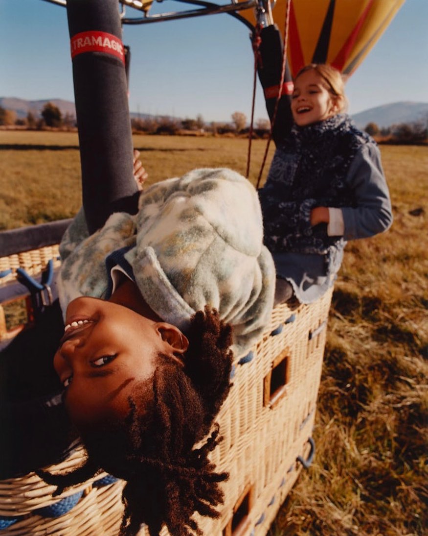 Two girls in a hot air balloon smile. One is upside down, facing the camera. The other is upright with her arm raised in the air.