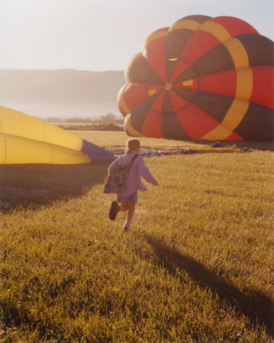 A child runs across a grassy field toward a partially deflated hot air balloon, with mountains visible in the hazy background at sunset.