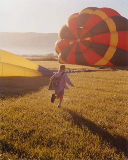 A child runs across a grassy field toward a partially deflated hot air balloon, with mountains visible in the hazy background at sunset.