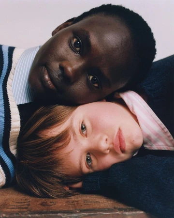 Close-up portrait of two young models with their heads resting together on a textured wooden surface against a neutral background.