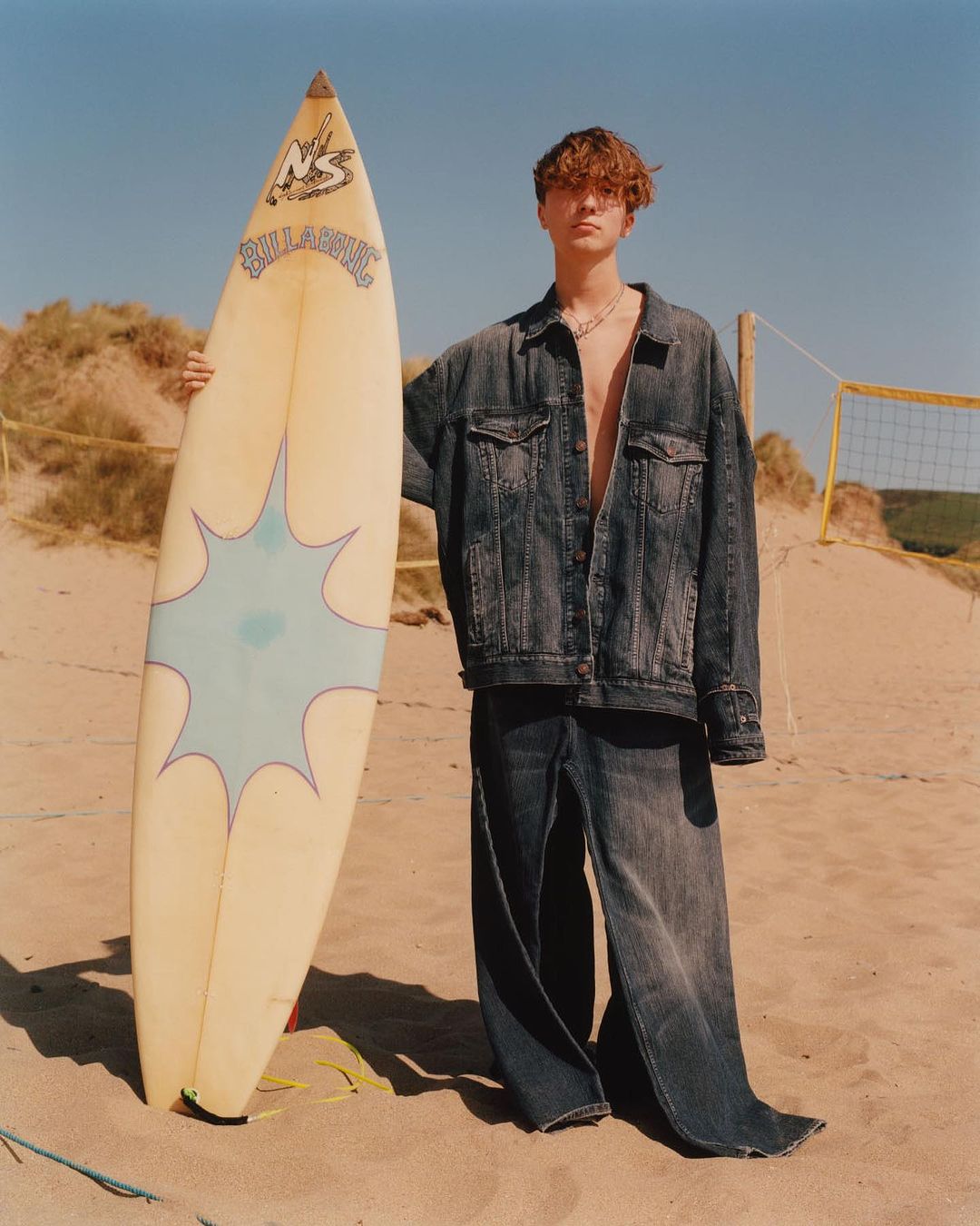Young man poses on a sandy beach in oversized denim outfit, holding a surfboard with vintage graphic art, under a clear blue sky.