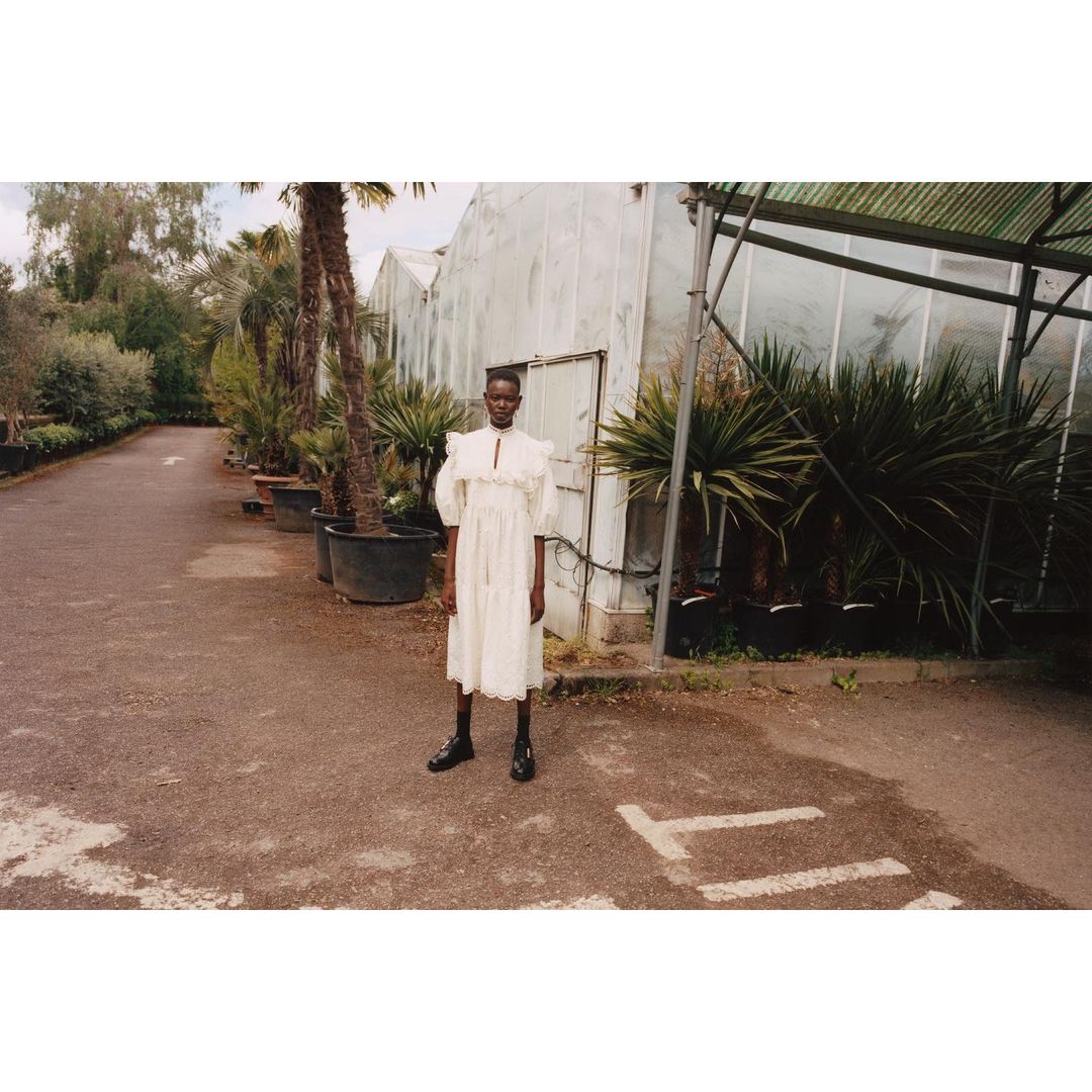 Woman in a white ruffled dress and black boots stands outside a greenhouse on a partially paved driveway, surrounded by potted plants.