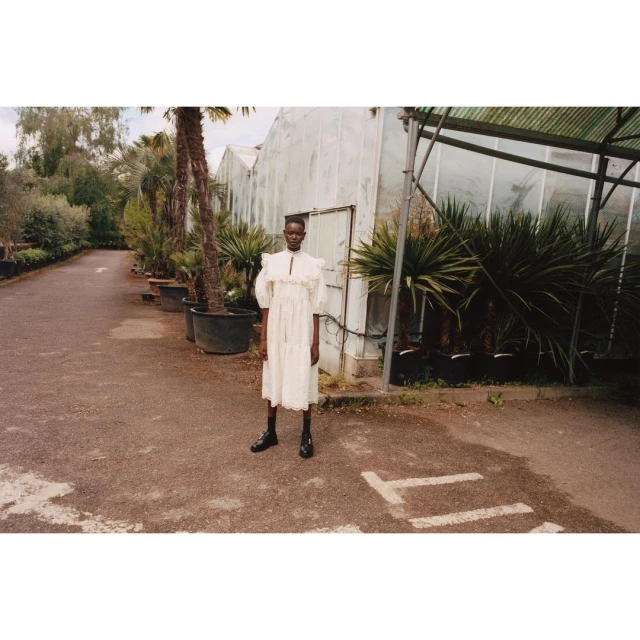 Woman in a white ruffled dress and black boots stands outside a greenhouse on a partially paved driveway, surrounded by potted plants.