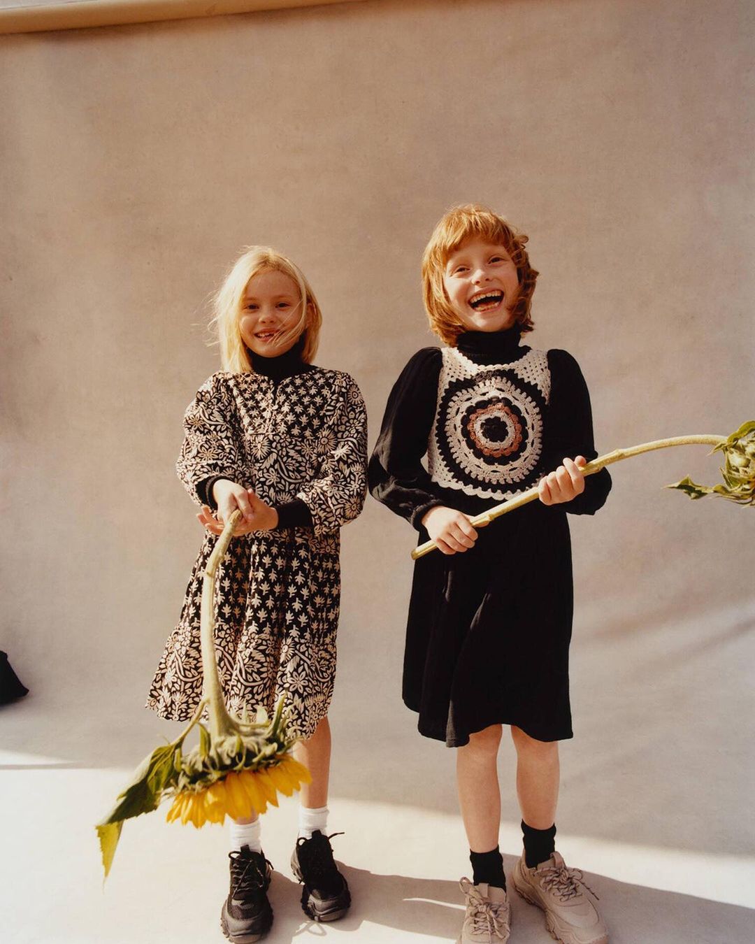 Two smiling girls in dresses pose with a sunflower against a neutral backdrop, captured in a medium shot with natural lighting.