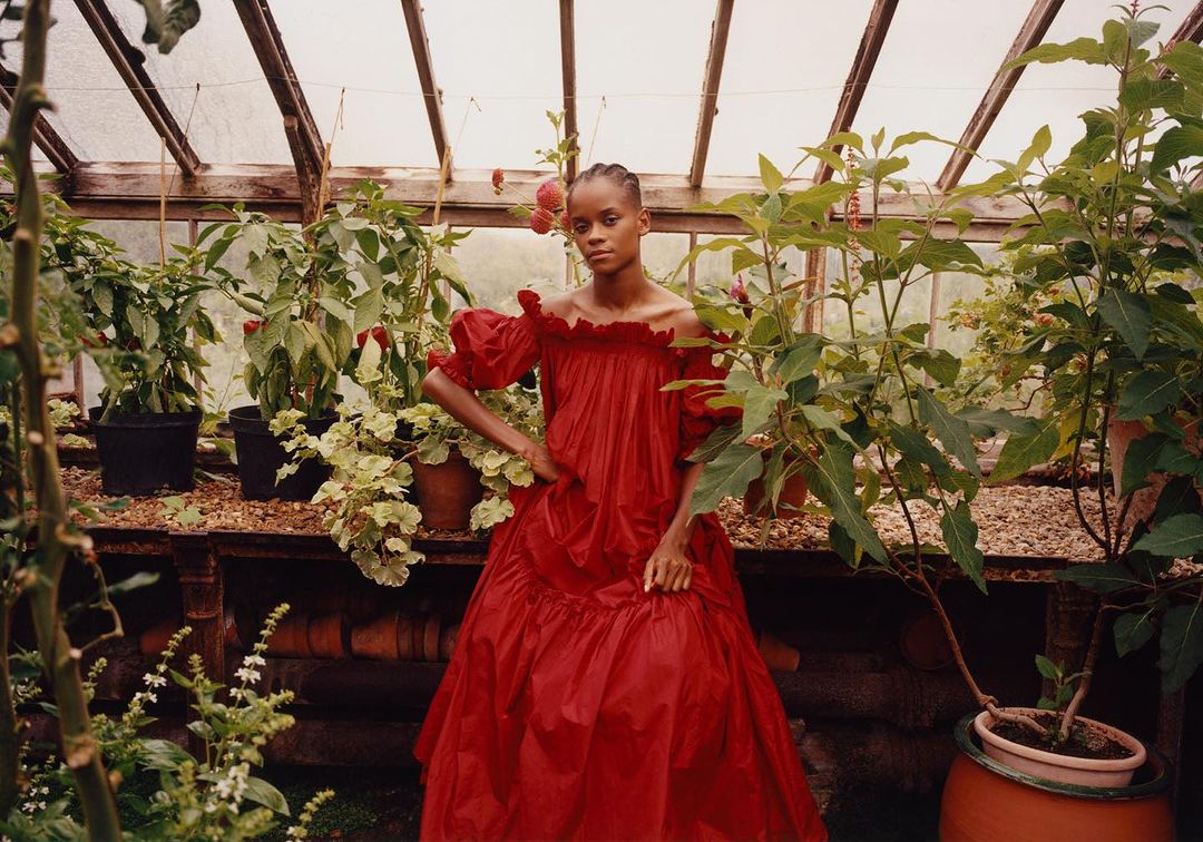 A woman in a red dress poses in a greenhouse filled with potted plants under a clear, angled roof. The light is diffused and natural.