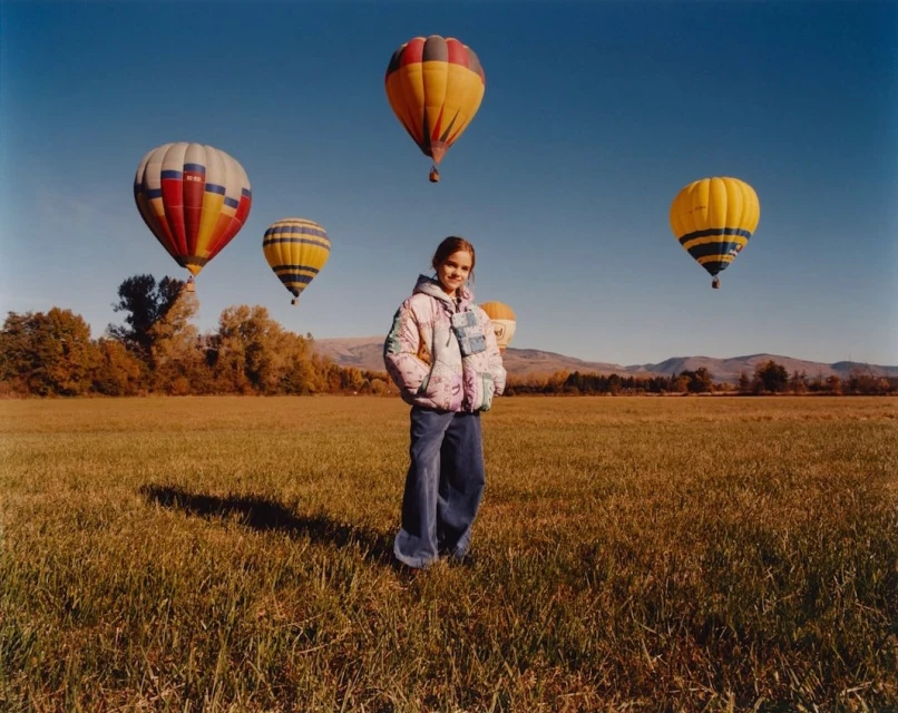 A young woman stands in a field with several colorful hot air balloons floating in the sky on a sunny day. A treeline is visible in the distance.