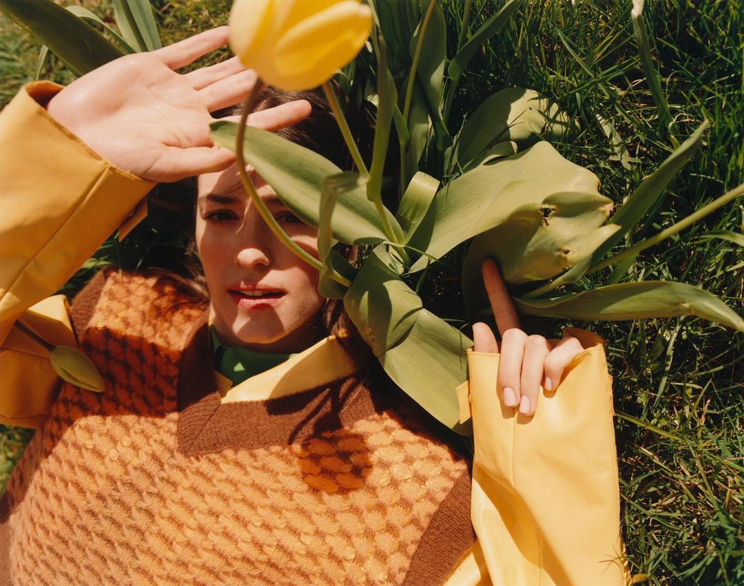 Woman lies in the grass in a yellow sweater, shielding her eyes from the sun with her hand, with a yellow tulip in foreground.