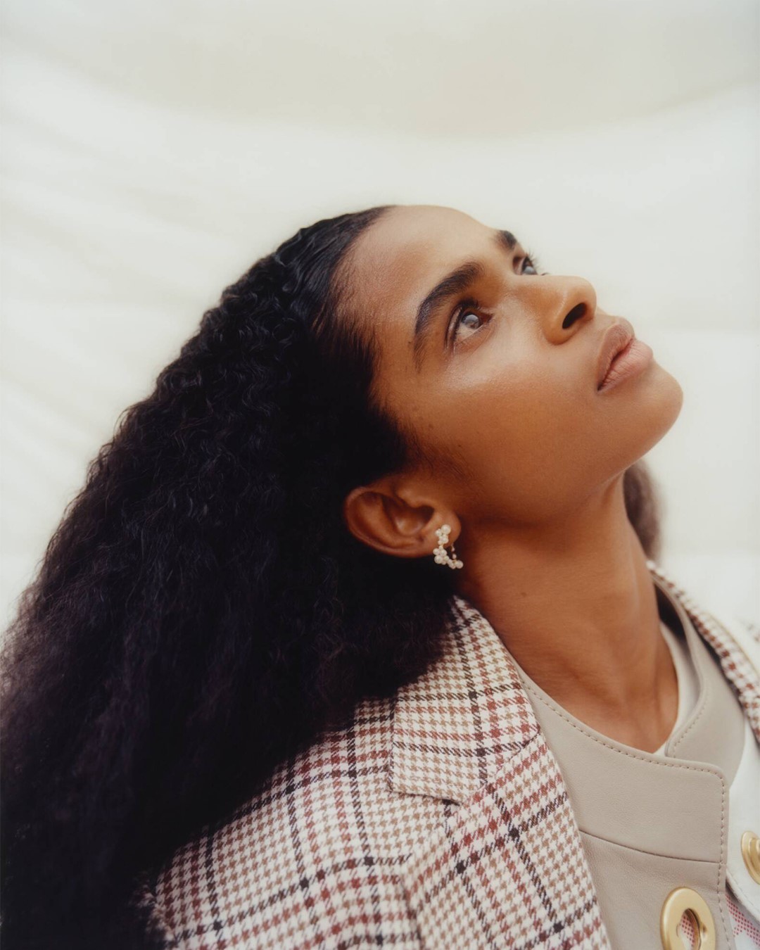 Portrait of an adult female model with curly black hair, wearing earrings and a plaid jacket, looking upward against a neutral background.