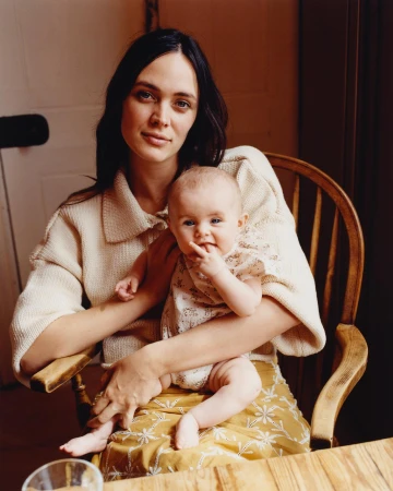 Mother and baby portrait. Adult woman with long dark hair, wearing a cream sweater, holds a baby on her lap in a wooden chair.