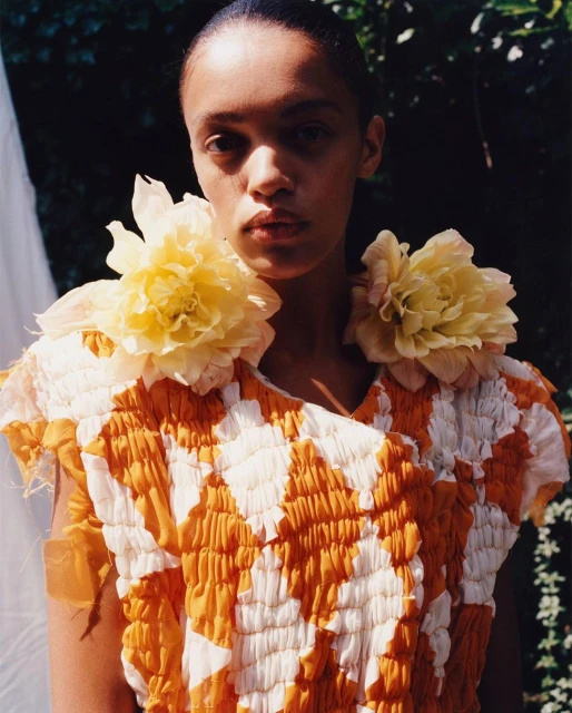 Portrait of an adult woman in a yellow and white patterned top with flower details, posing outdoors in natural lighting.
