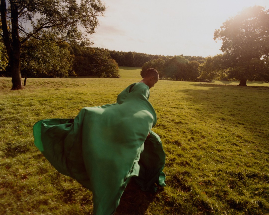 Woman in a flowing green gown walks through a sunny meadow with trees in the background, creating a serene and fashionable scene.