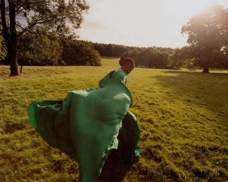 Woman in a flowing green gown walks through a sunny meadow with trees in the background, creating a serene and fashionable scene.