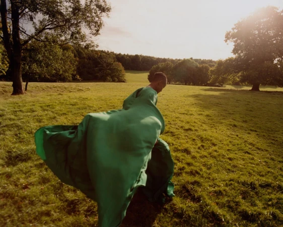 Woman in a flowing green gown walks through a sunny meadow with trees in the background, creating a serene and fashionable scene.