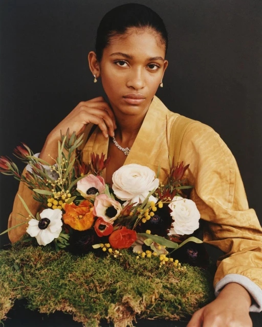 Elegant woman poses with a vibrant flower arrangement. She wears a yellow jacket and jewelry against a dark background in a portrait.