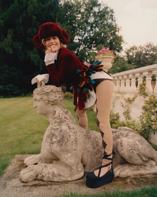 Woman in a costume leans on a stone sphynx in a garden. She's wearing a velvet coat, hat, platform shoes, and patterned leggings.