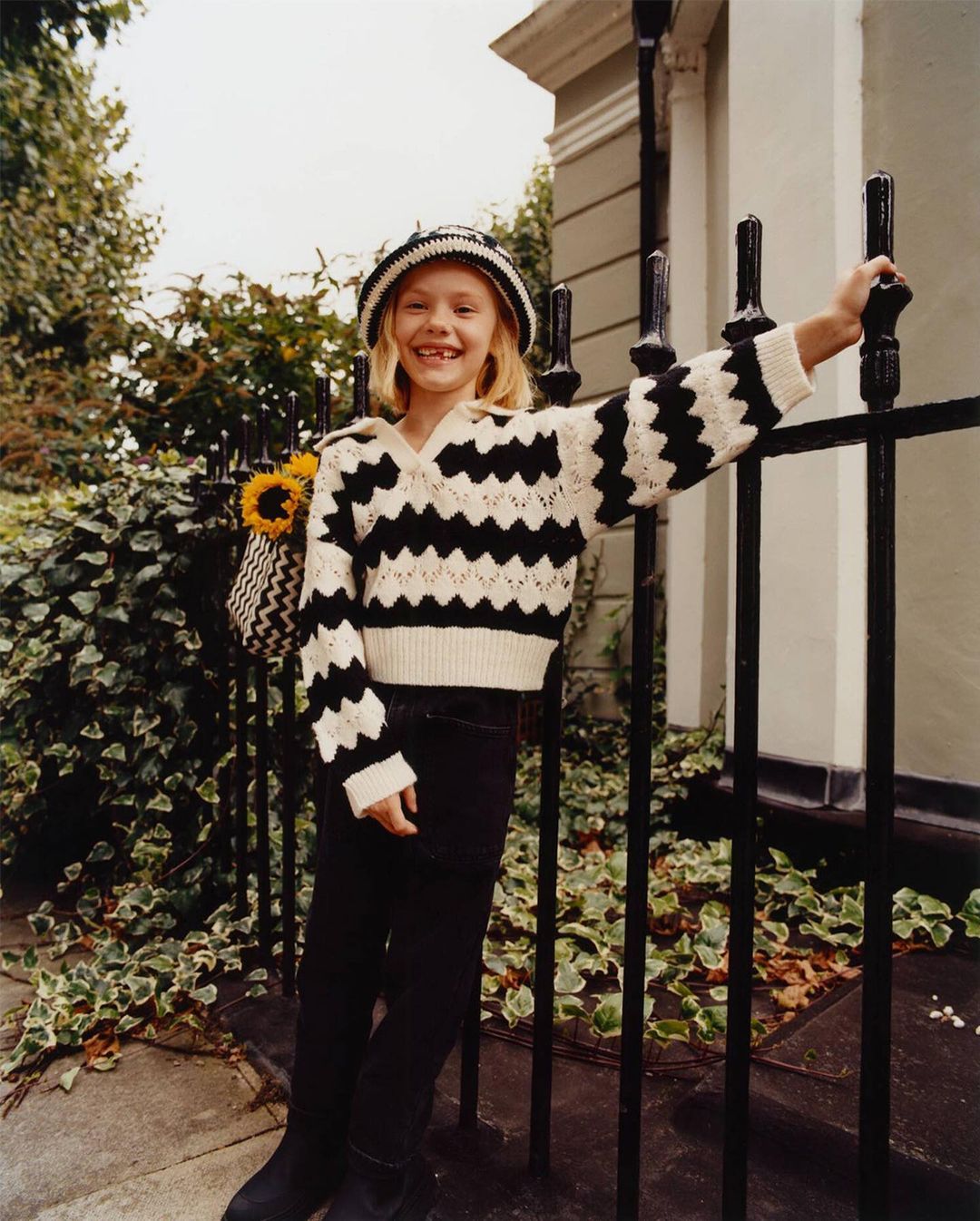 Child model smiles wearing a black and white zig-zag sweater and hat leaning against a black metal fence outdoors.