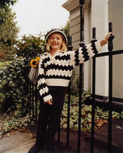 Child model smiles wearing a black and white zig-zag sweater and hat leaning against a black metal fence outdoors.