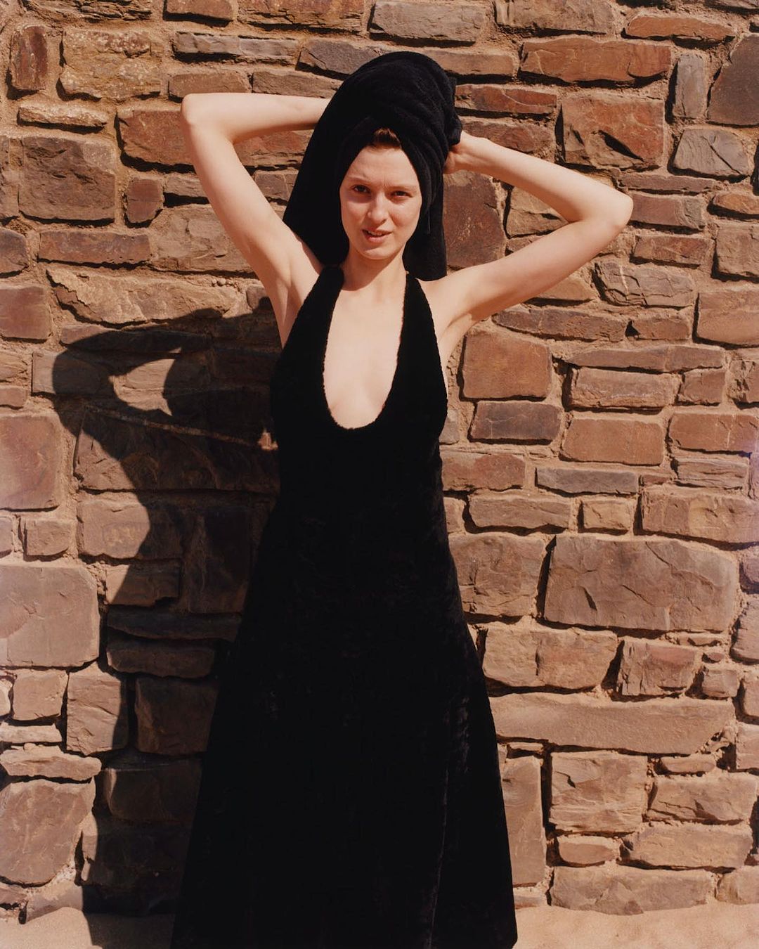 A young adult woman wearing a black dress and black towel on her head stands in front of a rustic stone wall in an outdoor setting.