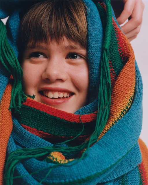 Close-up of a smiling child framed by a colorful striped scarf. The scarf features a mix of teal, green, red, and orange hues.