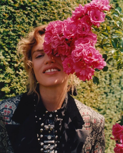 Close-up of a woman with blonde wavy hair, half her face obscured by a cluster of pink roses, wearing a patterned jacket outdoors.