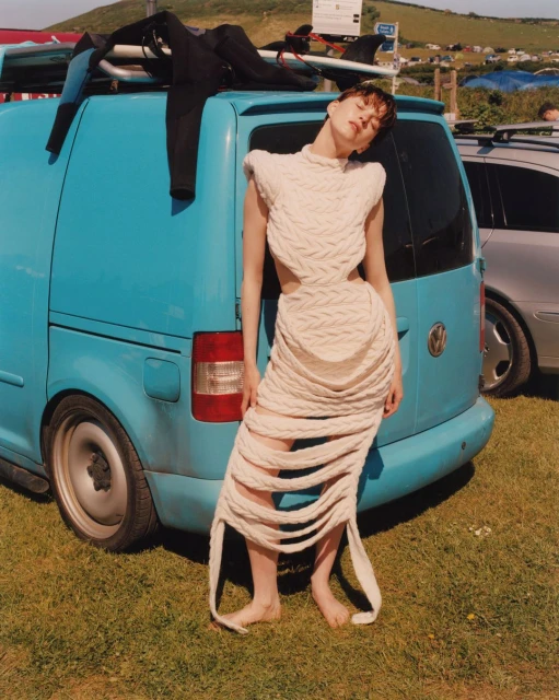 Fashion photo of a young woman in a knitted dress standing in front of a blue van with surfboards in a grassy outdoor setting.