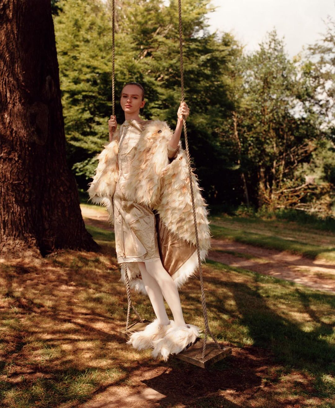 A young woman in a vintage-style feathered outfit poses on a wooden swing under a large tree in a sun-dappled forest setting.