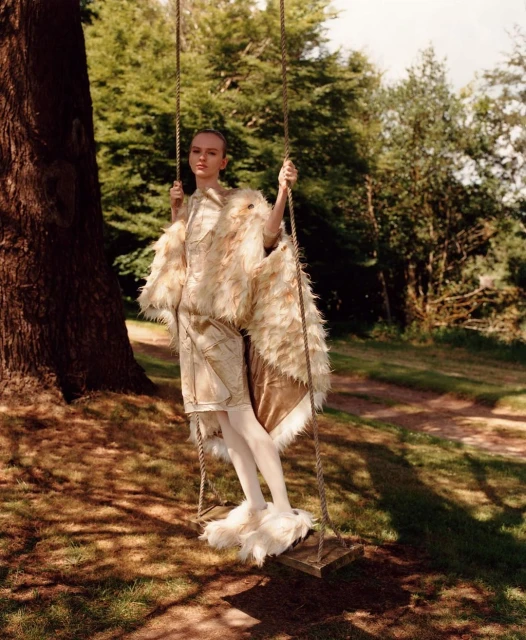 A young woman in a vintage-style feathered outfit poses on a wooden swing under a large tree in a sun-dappled forest setting.