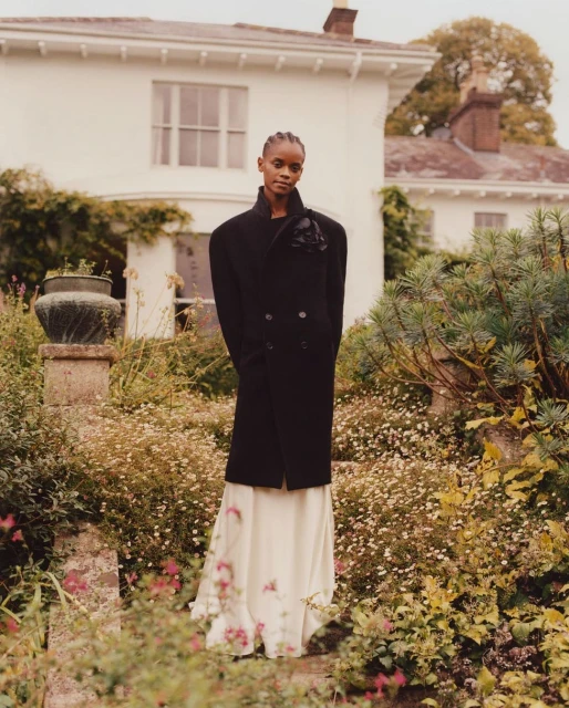 A woman in a black coat and a long white skirt poses in a lush garden in front of a white country house. The image has soft, natural light.