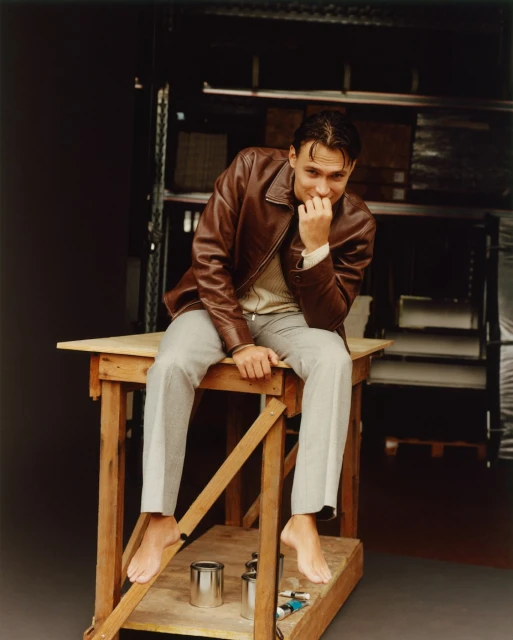 A barefoot adult male sits on a wooden structure in a brown leather jacket, light sweater, and grey slacks in an indoor setting.