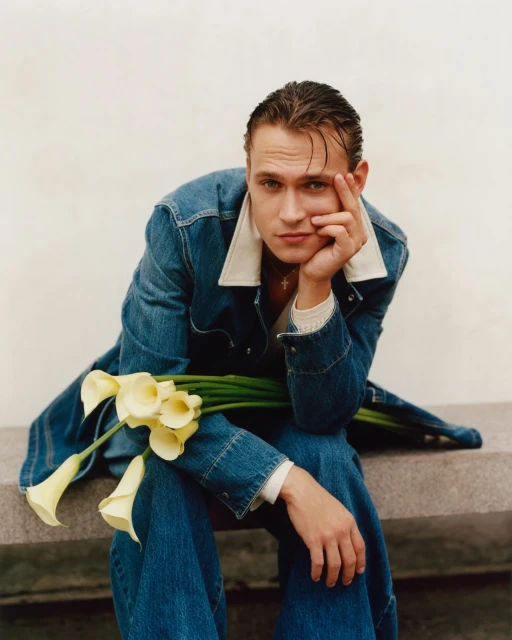 Pensive male model in denim outfit poses with calla lilies in a studio portrait, showcasing fashion and floral elements on a neutral backdrop.