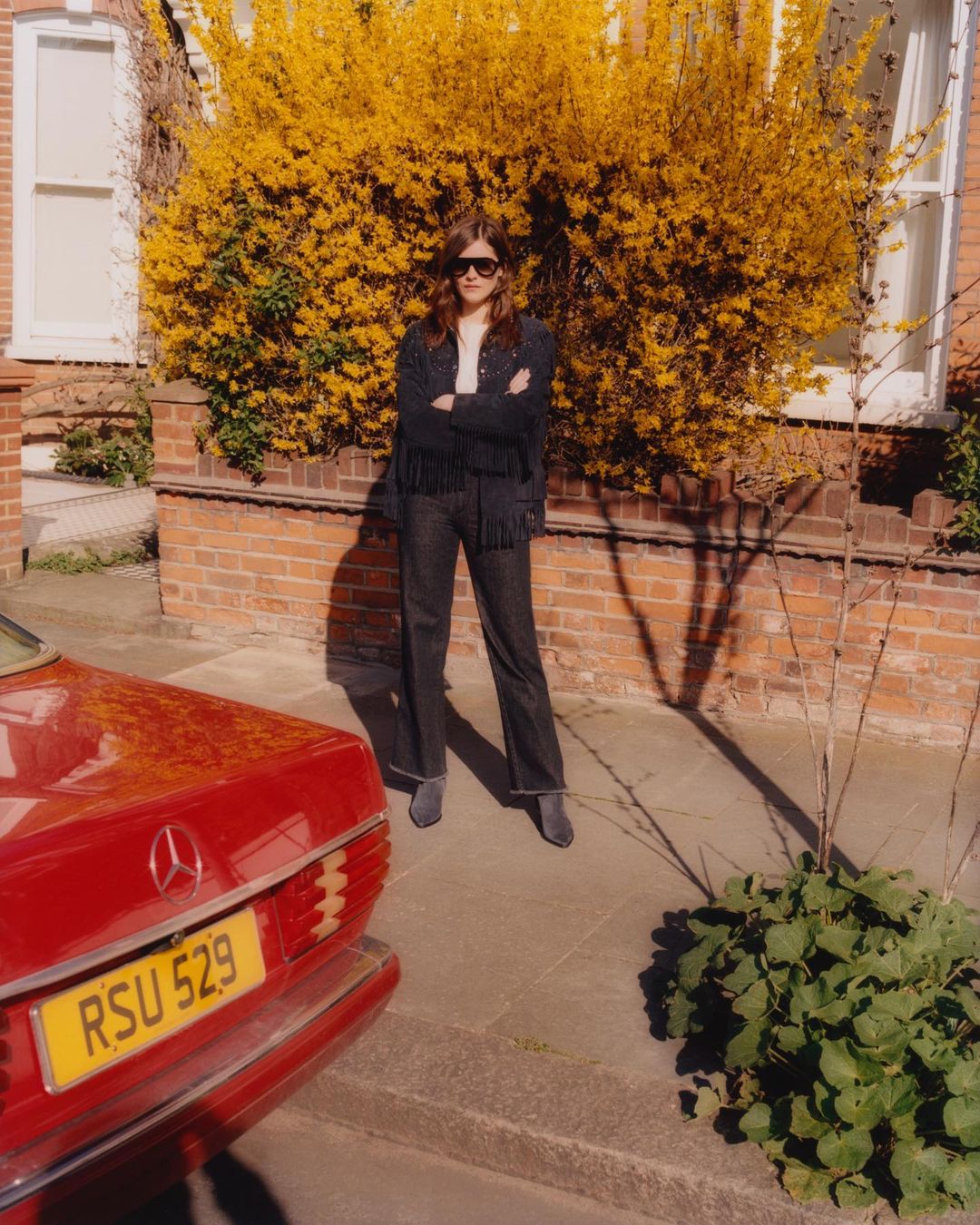 Woman poses in jacket with fringe next to vintage Mercedes-Benz car in residential neighborhood on a sunny day, vibrant yellow shrubbery.